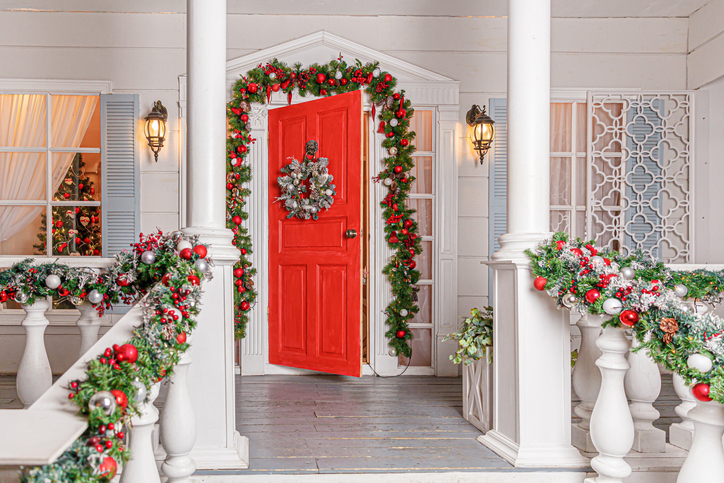 Christmas porch decoration idea. House entrance with red door decorated for holidays. Red and green wreath garland of fir tree branches and lights on railing. Christmas eve at home.