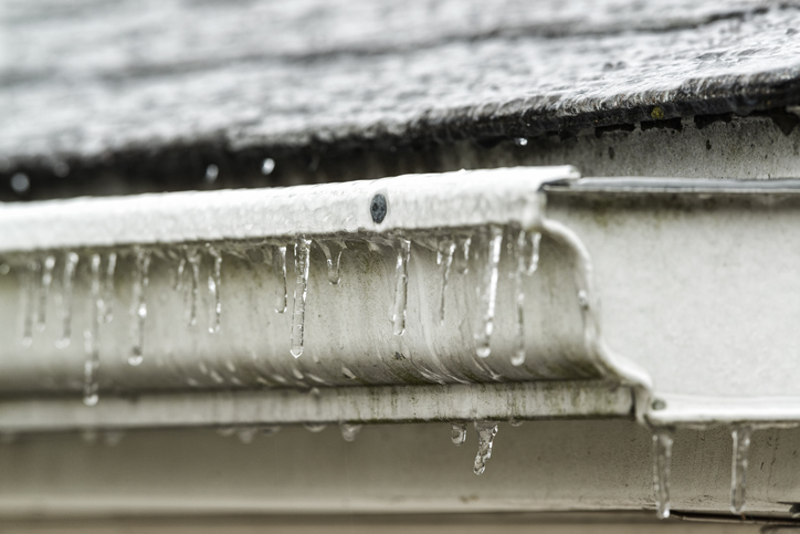 Close-up of icicles forming on an old white residential gutter from freezing rain drips off the shingled roof on a cold Winter day. Home owner's need to be careful of ice dams that can lead to roof leaks.