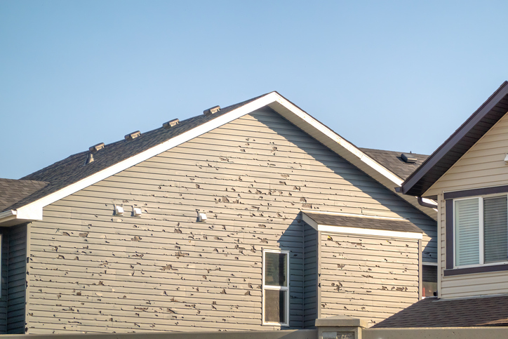 A residential home exhibits substantial hail damage to its vinyl siding, characterized by numerous small dents and chips.